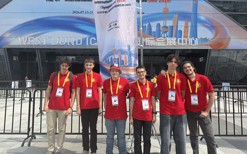 olimpiada-mate - Sursa de Vest Six young people in red shirts with lanyards pose for a photo outside a modern expo center with a tall blue banner in the background.