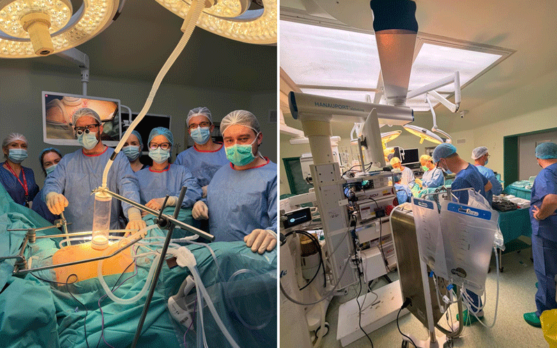 Surgeons and nurses in blue gowns and masks operate around a patient under bright overhead lights in an operating room.