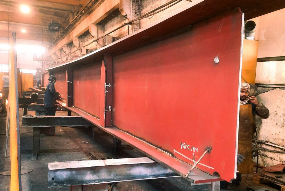 piese pod muncii2 - Sursa de Vest Welding workshop with a long red metal panel on a stand and a worker in protective gear welding it.