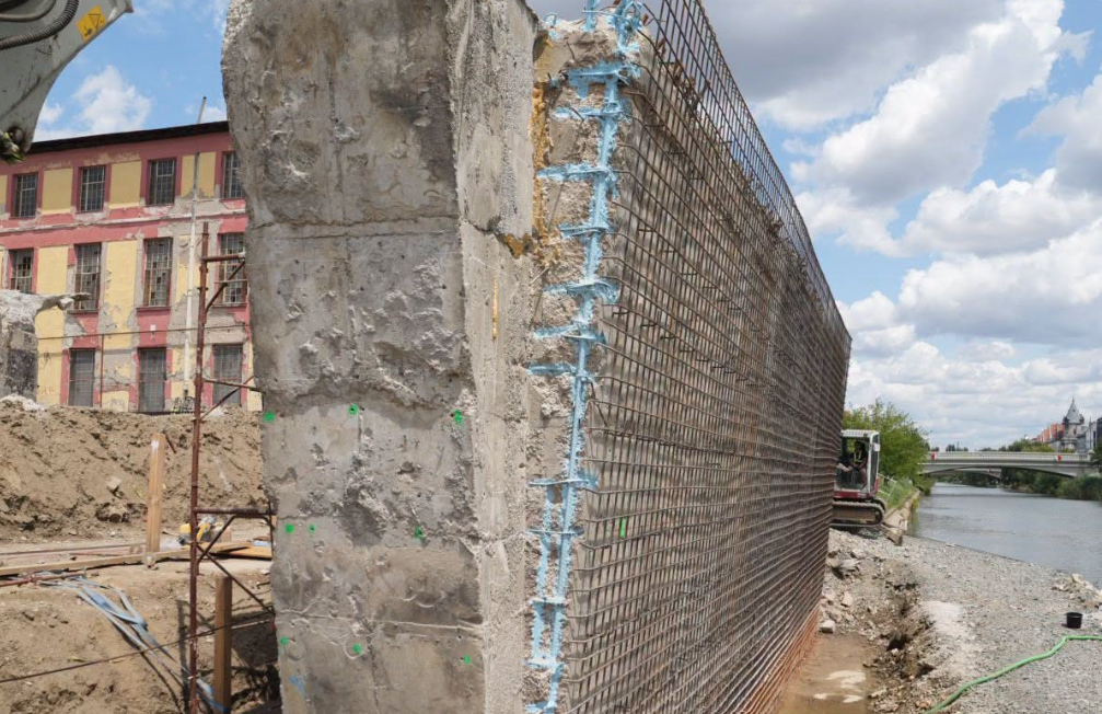 podul muncii2 - Sursa de Vest Construction site along a river with a tall steel-rebar wall and blue waterproofing, a digger arm visible nearby, and a colorful building in the background.