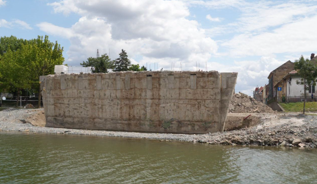 podul muncii3 - Sursa de Vest Large rectangular concrete barrier along a riverbank under construction, with rubble piles and trees in the background and houses beyond.