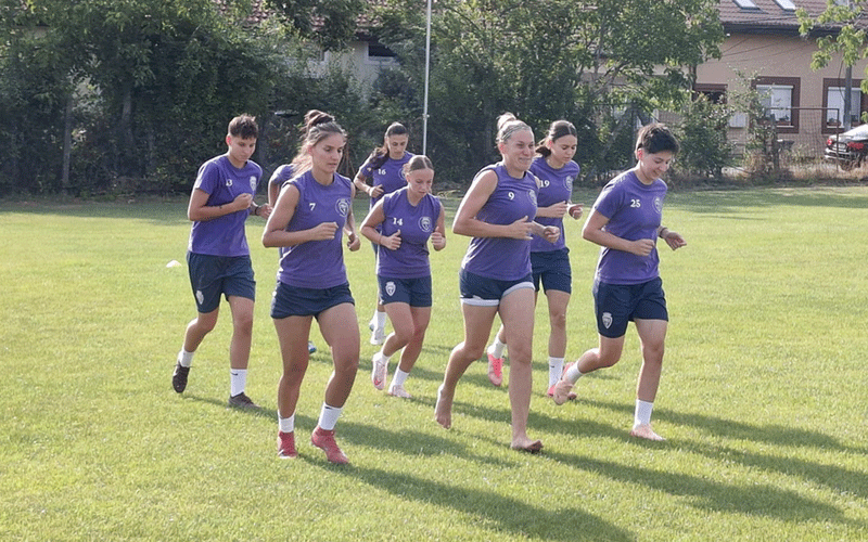 Group of young women in matching purple jerseys jogging together on a sunny grassy field, likely a team training session.