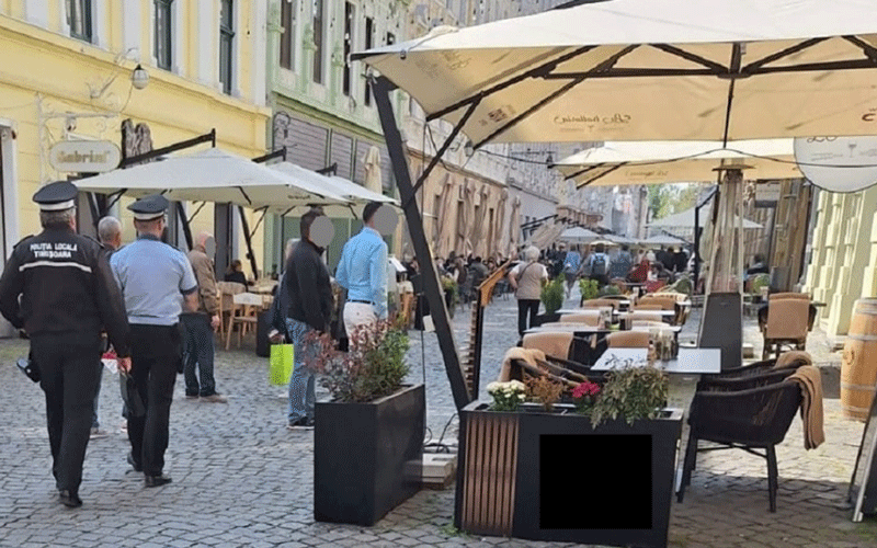 politia-locala - Sursa de Vest Outdoor cafe along a cobblestone street with white umbrellas, tables, and pedestrians.