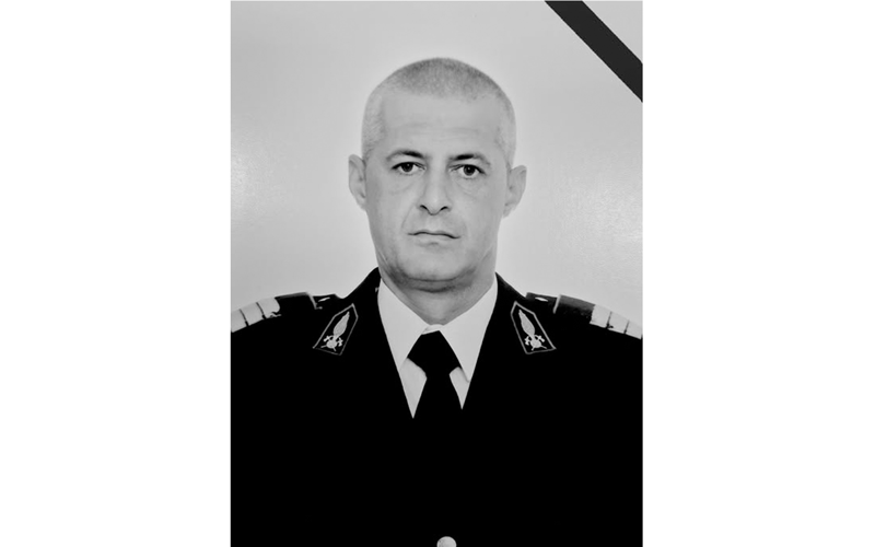 Portrait of a male officer in a formal dress uniform (black suit, tie) in grayscale, facing the camera with a neutral expression, mourning ribbon in the top-right corner.