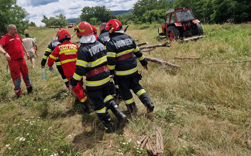 pompieri-tractor - Sursa de Vest Firefighters in black turnout gear walk through a sunlit grassy field, with a red tractor in the background.