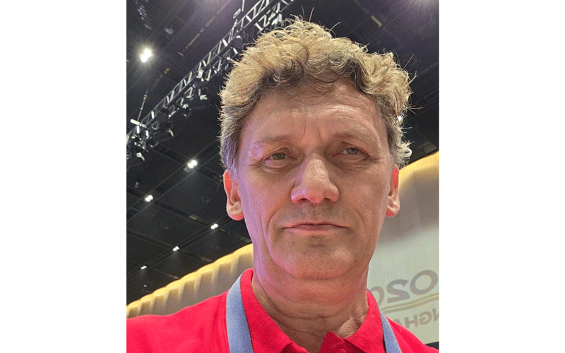 Portrait of a middle-aged man with wavy light brown hair wearing a red polo and a blue lanyard at an indoor event venue