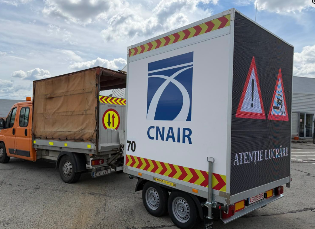 remorca drdp - Sursa de Vest Truck with a large white trailer displaying the CNAIR logo, attached to an orange cab, on a paved lot with a blue-sky background and safety chevrons along the edges.”,
