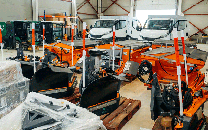 Indoor warehouse displaying orange snow plows and white delivery vans in the background, with pallets and equipment nearby.