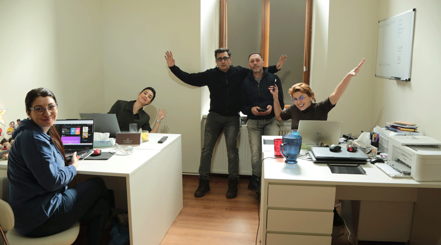 Five coworkers in a bright office celebrate, two seated at white desks with laptops and three standing in back with arms raised in excitement.