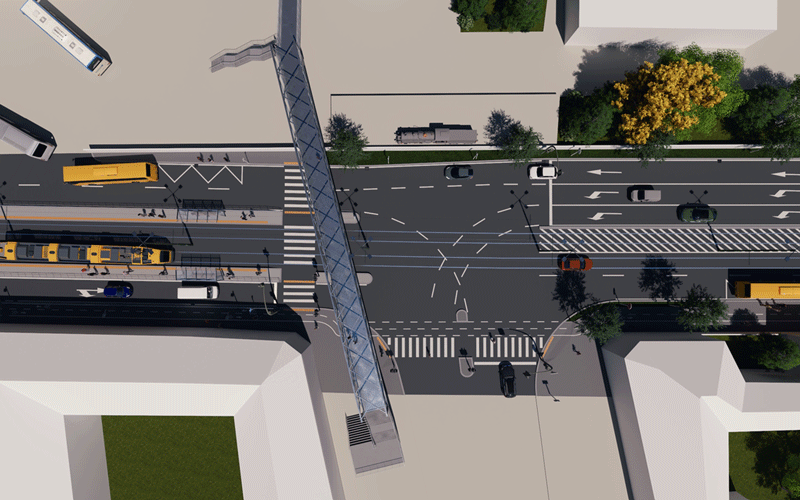 Aerial view of a busy city intersection with a glass pedestrian bridge crossing the road, buses and cars on multi-lane streets, crosswalks, and surrounding buildings and trees.