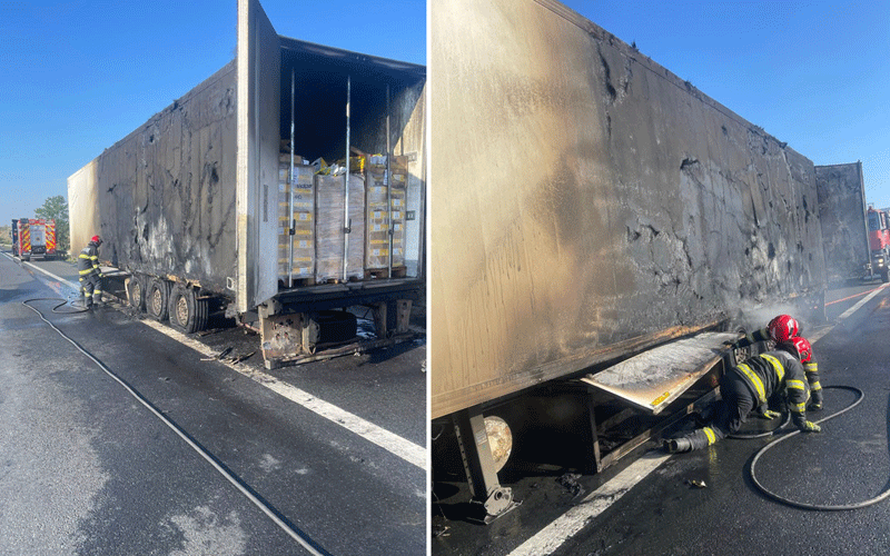 Burnt semi-trailer on a highway with firefighters spraying water on the damaged side and cargo pallets exposed inside the trailer.