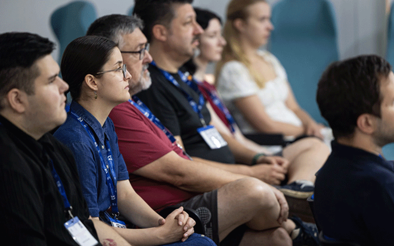 Several diverse people seated in a conference room, wearing lanyards and listening attentively to a presentation.