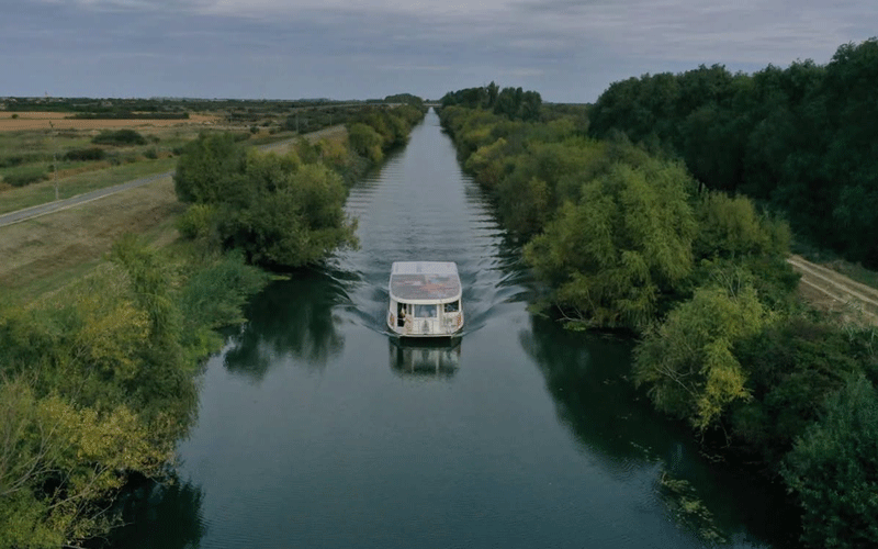 A boat cruising down a narrow canal lined with green trees on both sides, viewed from above.