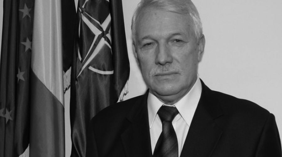 Grey-haired man in a suit and tie posing for a formal portrait with flags in the background.