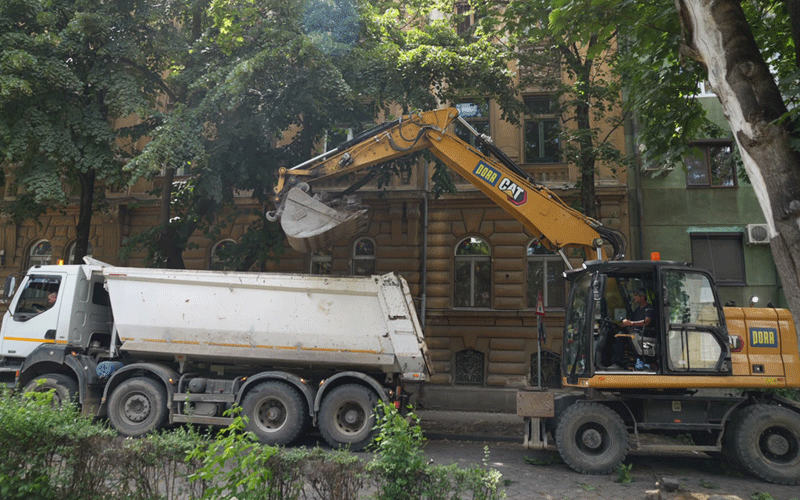 zona-traian - Sursa de Vest Yellow excavator loading soil into a white dump truck on a city street, trees and a historic building in the background