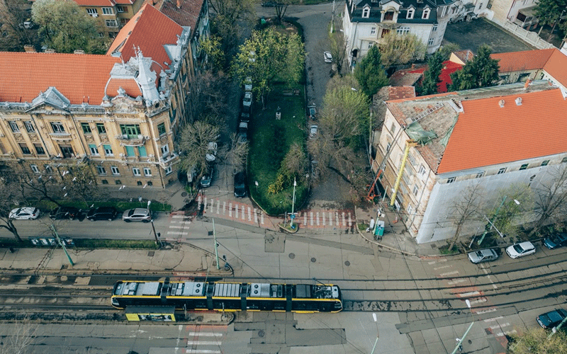 zona-traian2 - Sursa de Vest Aerial view of a tram moving along a city street between red-roofed buildings and a tree-lined median.