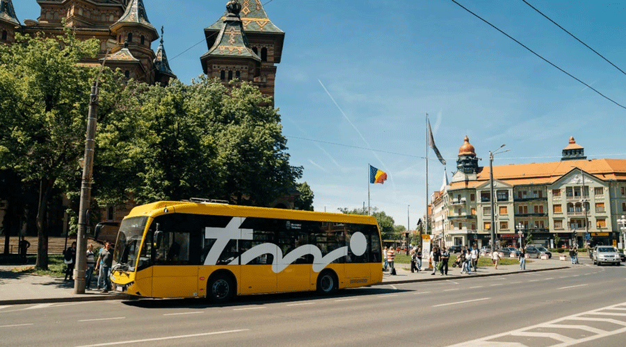 Yellow tour bus parked on a city street with pedestrians on the sidewalk and historic buildings in the background, Romania flag visible above.