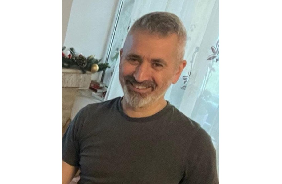 Smiling man with short gray hair and a gray beard, wearing a dark t-shirt, indoors by a window.