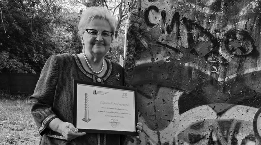Older woman with short light hair and glasses, wearing a pearl necklace, holds a framed diploma outdoors beside a graffiti-covered wall.