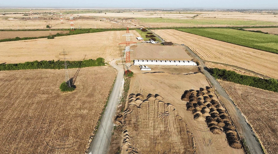 Aerial view of rural farmland with dirt roads, rows of hay bales, and a long white barn-like building near red transmission towers.