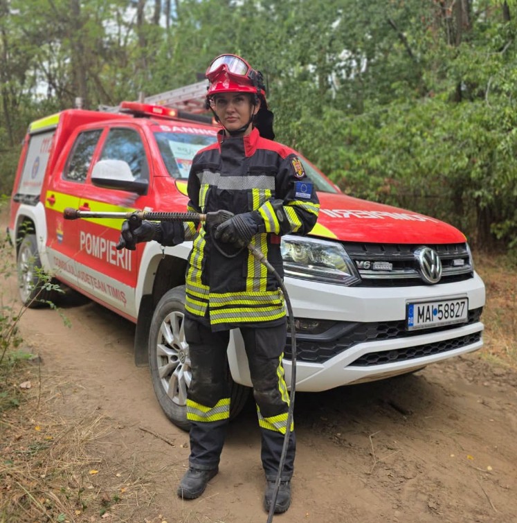 Firefighter in full turnout gear holding a hose, standing in front of a red Romanian fire truck on a dirt road in a forested area.
