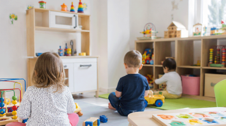 Three children play with toys in a bright classroom with shelves and a pretend kitchen.
