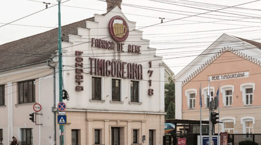 Historic street scene with a white, stepped-gable factory building reading 'Fabrica de Pere' and large signage; overhead wires cross the street in front of pastel-colored buildings.