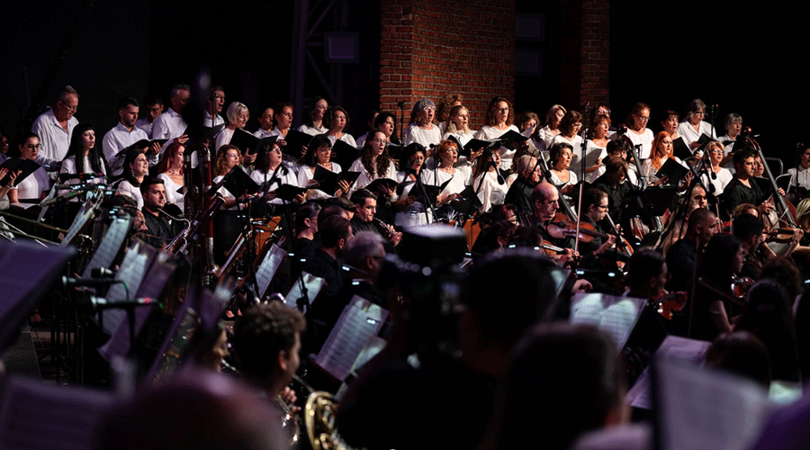 Large choir standing behind an orchestra on a dimly lit stage during a performance.