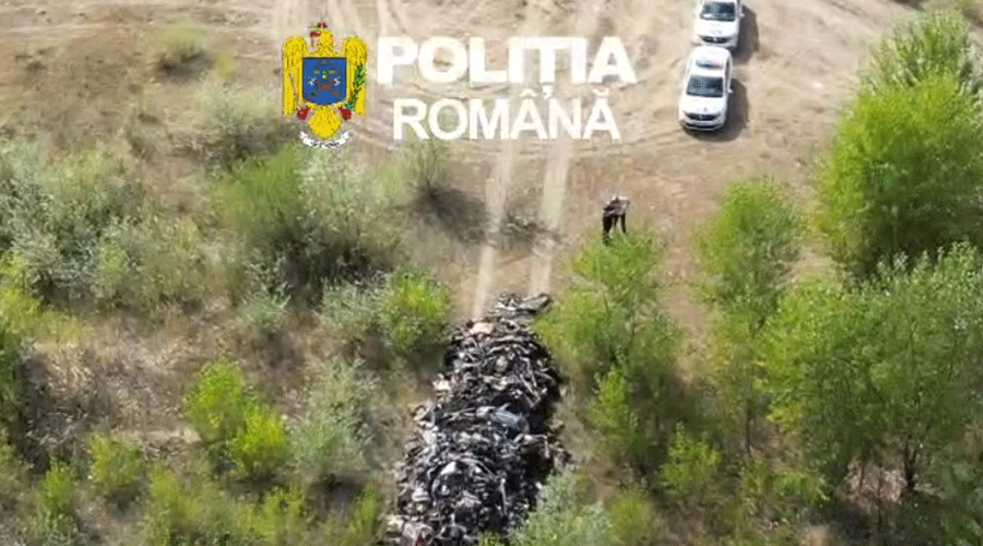 Aerial view of a long pile of body bags along a dirt path in a forest, with police cars nearby and the Romanian Police emblem visible in the corner.