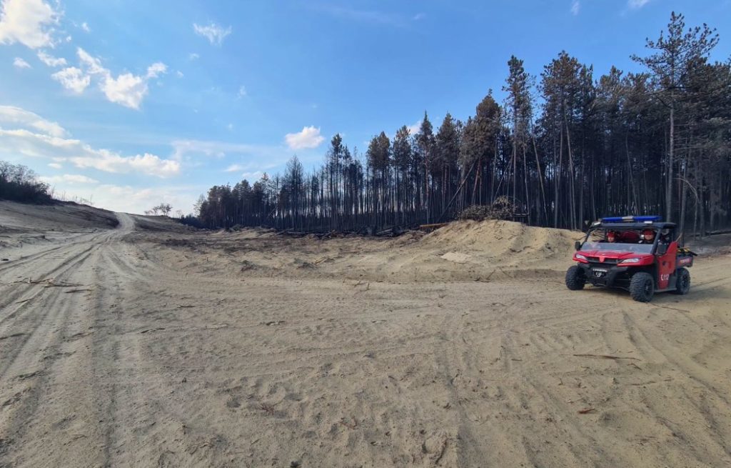 Red off-road utility vehicle parked on a sandy clearing with a line of burnt trees and a dirt mound in the background.