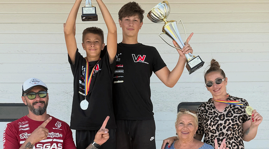 Two young boys stand on a podium holding trophies, medals around their necks, flanked by adults celebrating the win.
