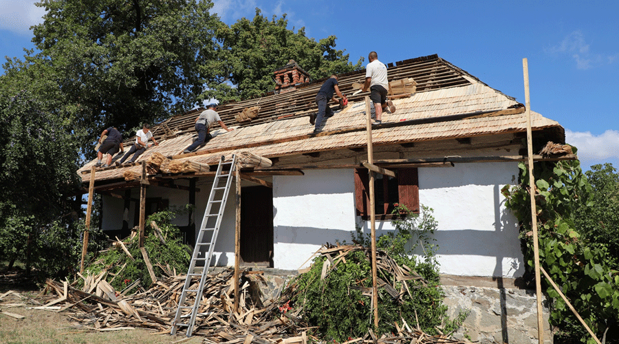 Workers repair a thatched roof atop a white cottage, using ladders and scaffolding outdoors.