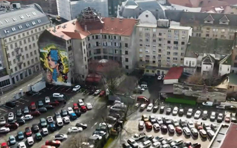 Aerial view of a European city square with numerous parked cars and pink-beige buildings, plus a large mural of a woman on the left wall.