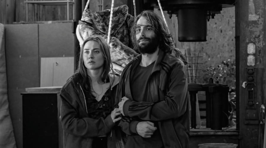 Two adults stand side by side in a dim, industrial workshop, looking ahead with tense expressions.