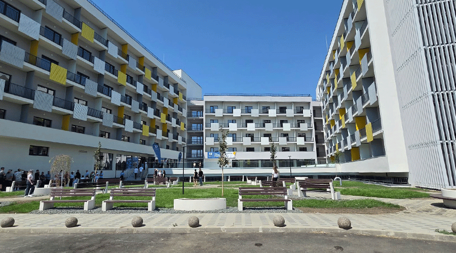 University campus courtyard with modern white buildings, yellow accents, benches, and people walking under a blue sky.