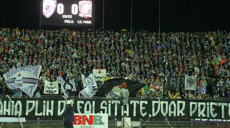 Night-game crowd of football supporters in a stadium, waving banners and flags, with a long banner spanning the front row.