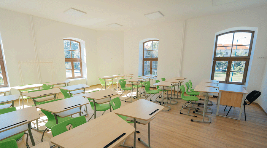 Bright empty classroom with light wood desks and green chairs arranged in rows, large arched windows letting in natural light.