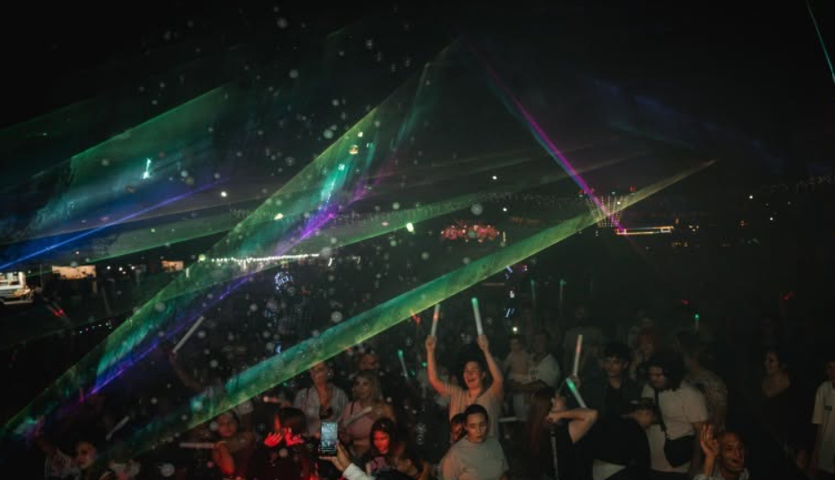 Crowd at a nighttime concert waving glow sticks as green and colorful laser beams cross overhead.