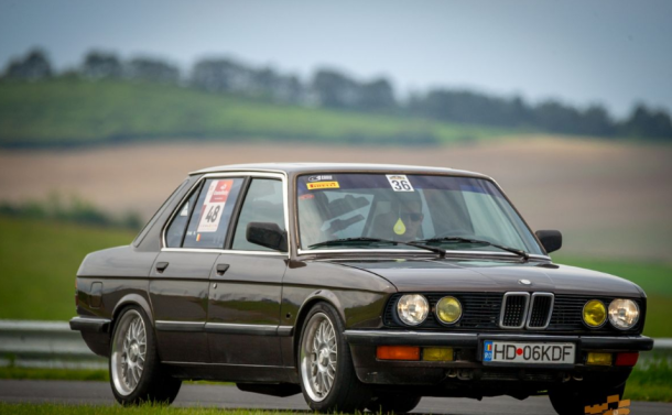 Brown 1980s BMW race car with numbered stickers on a track, front three-quarter view.