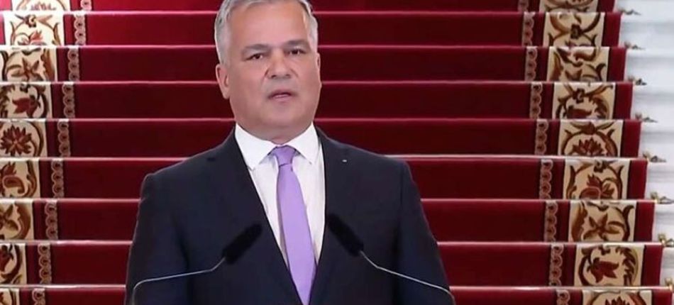Man in a dark suit and lavender tie delivering a speech at a podium, with red-carpet stairs behind him.