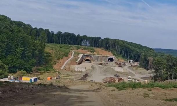 Construction site on a hillside with a tunnel entrance under an embankment, dirt roads and equipment nearby