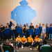 Group of people on a stage posing with blue jackets, in front of a large projected image of a blue hoodie on the screen behind them, an event photo showing staff and speakers holding up jackets.