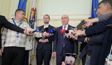 Bald man in a suit addressing reporters with microphones at a press conference, journalists surrounding him.
