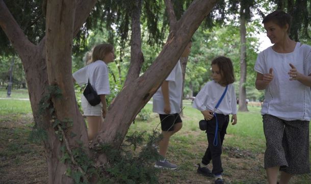 A woman and three children walk through a shaded park among tall trees, chatting as they go.