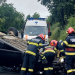 Emergency responders work at a single-vehicle crash with an overturned car on a rural road, ambulance in background.