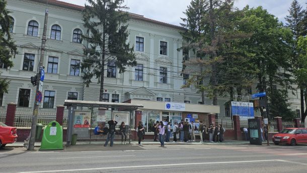 People wait at a bus stop in front of a pale green historic building with tall arched windows and trees nearby.