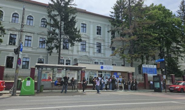 People wait at a bus stop in front of a pale green historic building with tall arched windows and trees nearby.
