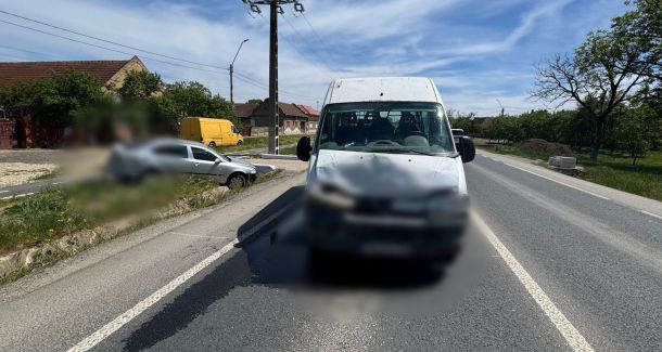White van in the foreground on a two-lane rural road after an accident; blurred front bumper and a silver car off the left roadside, blue sky above.