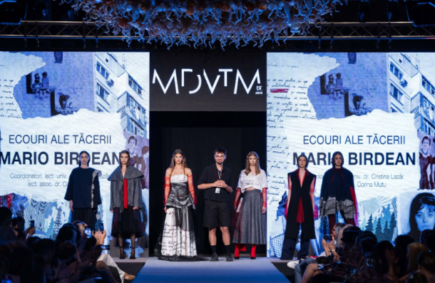 Runway fashion show: models in diverse outfits stand on stage under blue lighting; audience watches, with large screens showing text and images behind them.