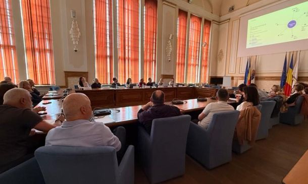 Formal meeting in a council chamber with people seated around a U-shaped wooden table and a large projector screen at the front.